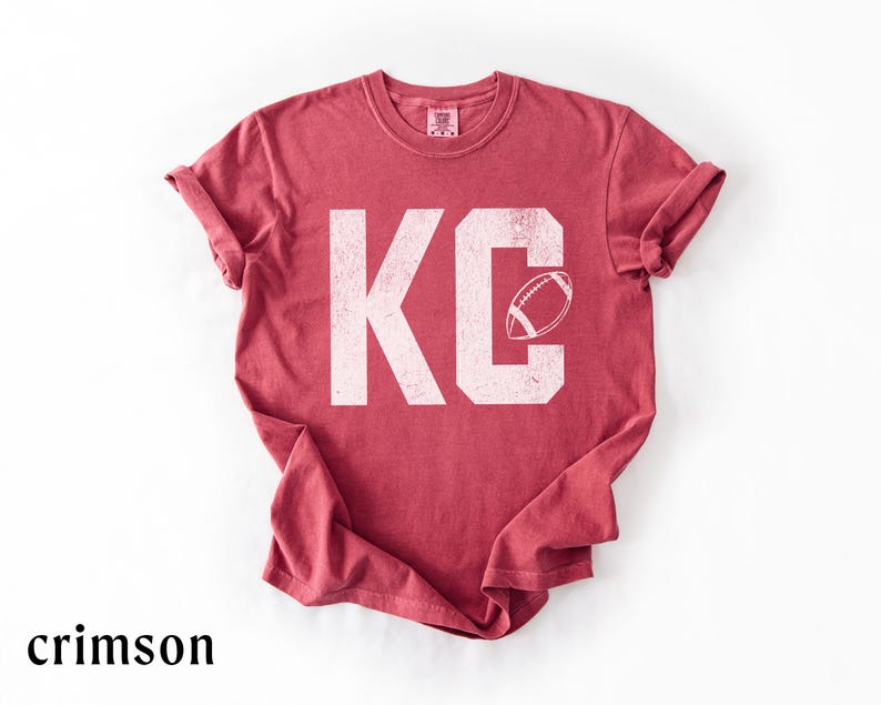 May include: Crimson-colored t-shirt with the letters "KC" in white, featuring a football graphic. The shirt has rolled-up sleeves and a crew neck. The word "crimson" is printed at the bottom of the image.