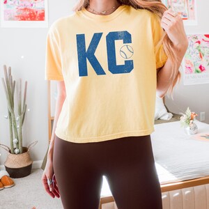 May include: A pale yellow t-shirt featuring the blue letters "KC" and a baseball graphic. The shirt has a crew neck and short sleeves. The word "butter" is visible in the bottom left corner.