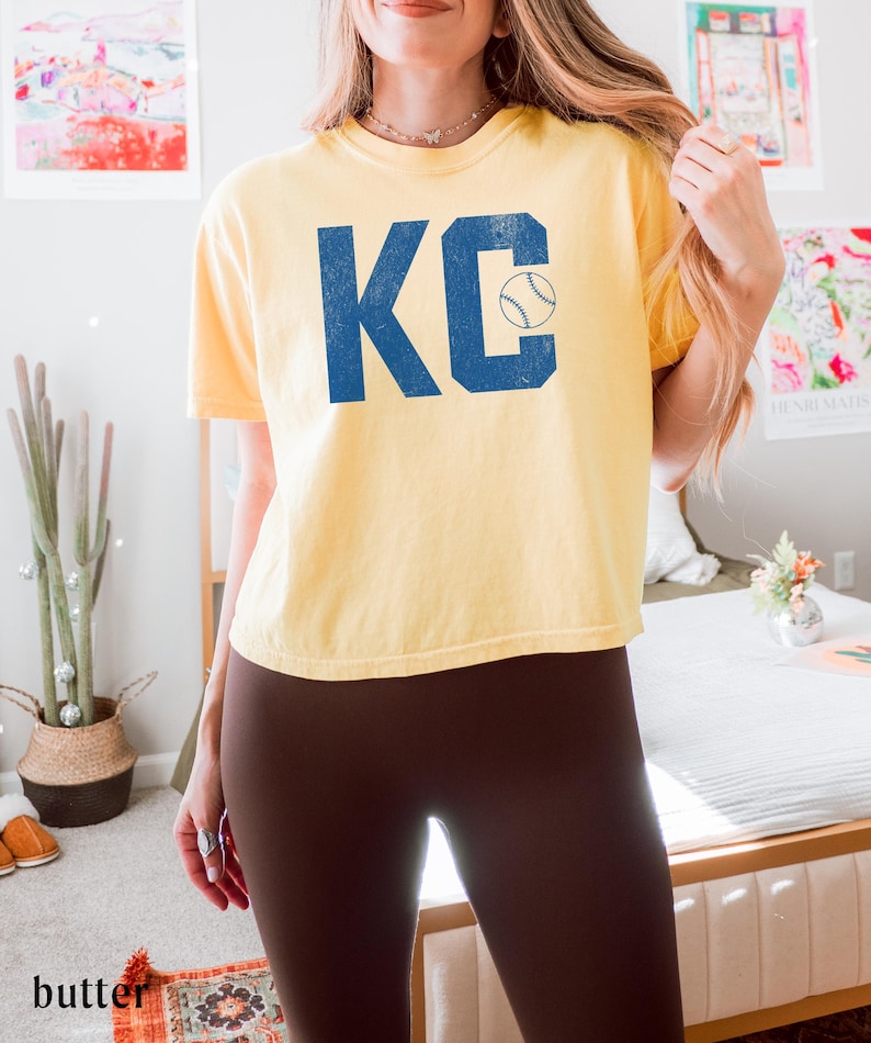 May include: A pale yellow t-shirt featuring the blue letters "KC" and a baseball graphic. The shirt has a crew neck and short sleeves. The word "butter" is visible in the bottom left corner.