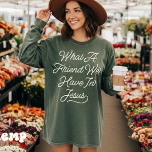 Könnte beinhalten: Ein olivgrünes Langarmshirt mit dem weißen Schriftzug "What A Friend We Have In Jesus". Die Person trägt eine Kaffeetasse. Im Hintergrund ein Blumenmarkt.
