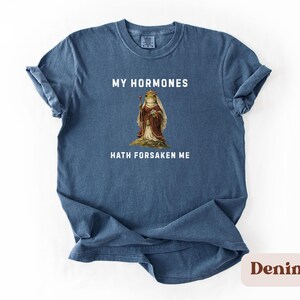 May include: A denim blue t-shirt featuring the phrase "MY HORMONES HATH FORSAKEN ME" above and below a vintage illustration of a frog in royal clothing. A casual wear item.