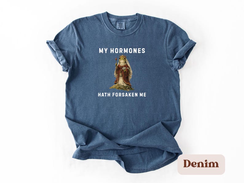 May include: A denim blue t-shirt featuring the phrase "MY HORMONES HATH FORSAKEN ME" above and below a vintage illustration of a frog in royal clothing. A casual wear item.