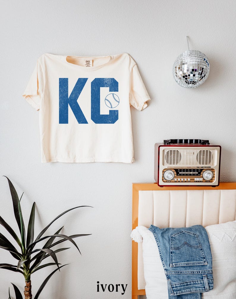May include: An ivory t-shirt with the blue letters "KC" and a baseball graphic. The shirt is hanging on a white wall. A disco ball, a vintage radio, and a plant are also visible in the image.