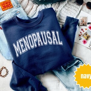 May include: Navy blue sweatshirt with the word "MENOPAUSAL" in white, collegiate-style lettering. The sweatshirt is folded on top of a pair of light blue jeans. A gold-colored bracelet and a pair of sunglasses are also visible.