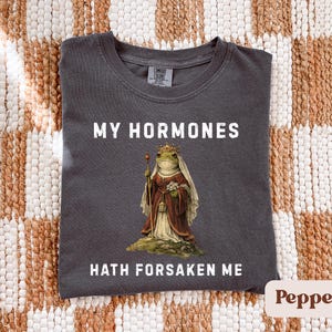 May include: Dark grey t-shirt with the text "MY HORMONES HATH FORSAKEN ME." The shirt features a regal frog illustration wearing a crown and holding a sceptre. The shirt is made of a soft material.