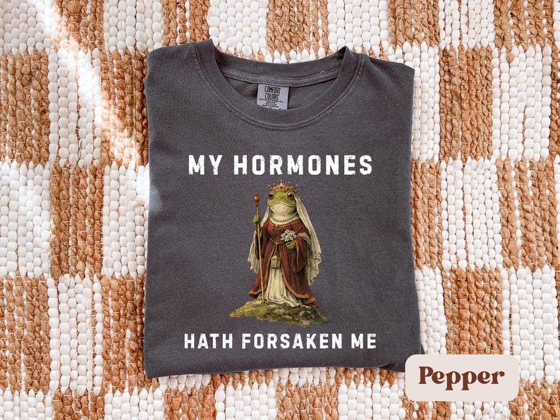 May include: Dark grey t-shirt with the text "MY HORMONES HATH FORSAKEN ME." The shirt features a regal frog illustration wearing a crown and holding a sceptre. The shirt is made of a soft material.