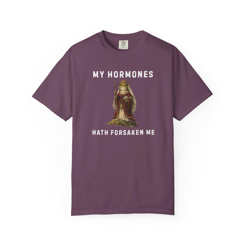 May include: A plum-coloured t-shirt with the text "MY HORMONES HATH FORSAKEN ME" in white. The graphic features a frog in regal attire. The shirt is a comfortable, casual wear item.