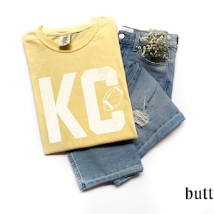 May include: A butter-yellow t-shirt with the letters "KC" and a football graphic, paired with light-wash denim jeans. The jeans have distressed details and a small bouquet of white flowers in the pocket. The word "butter" is at the bottom.