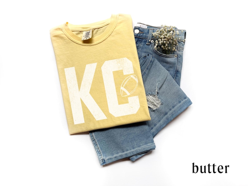 May include: A butter-yellow t-shirt with the letters "KC" and a football graphic, paired with light-wash denim jeans. The jeans have distressed details and a small bouquet of white flowers in the pocket. The word "butter" is at the bottom.