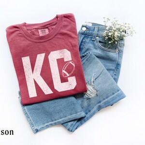 May include: Crimson-colored t-shirt with the letters "KC" and a football graphic, paired with light blue denim jeans. The t-shirt has a distressed look. A small bouquet of white flowers is tucked into the jeans pocket.