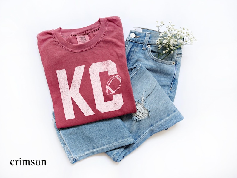May include: Crimson-colored t-shirt with the letters "KC" and a football graphic, paired with light blue denim jeans. The t-shirt has a distressed look. A small bouquet of white flowers is tucked into the jeans pocket.