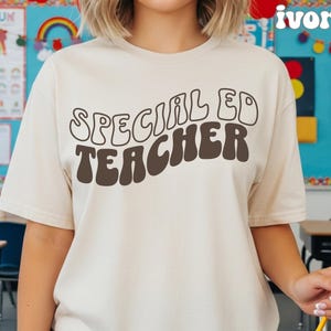Op de afbeelding: Een ivoorkleurig T-shirt met korte mouwen en de donkerbruine tekst "SPECIAL ED TEACHER" in een golvend, retro-geïnspireerd lettertype op de borst. Het shirt heeft een ronde hals en een standaard pasvorm met duidelijk gedrukte, ronde letters.