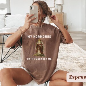 May include: Espresso-coloured t-shirt with the text "MY HORMONES HATH FORSAKEN ME" and a graphic of a frog in a robe. The shirt is a relaxed fit.