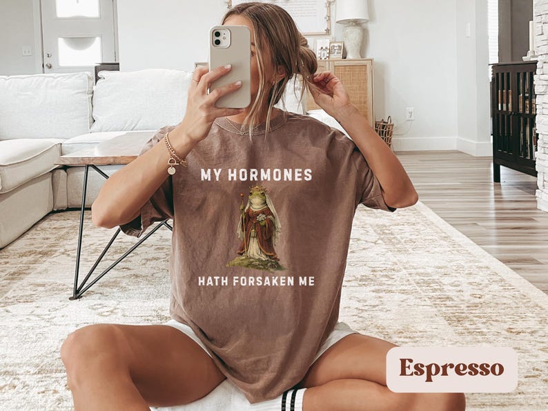 May include: Espresso-coloured t-shirt with the text "MY HORMONES HATH FORSAKEN ME" and a graphic of a frog in a robe. The shirt is a relaxed fit.