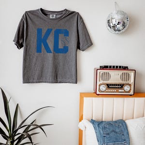 May include: A grey t-shirt featuring the blue letters "KC" and a baseball graphic. The shirt is hanging on a white wall. A disco ball, a vintage radio, and a pair of jeans are also visible.