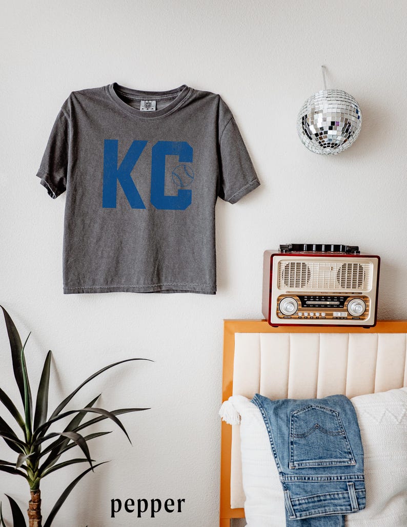 May include: A grey t-shirt featuring the blue letters "KC" and a baseball graphic. The shirt is hanging on a white wall. A disco ball, a vintage radio, and a pair of jeans are also visible.