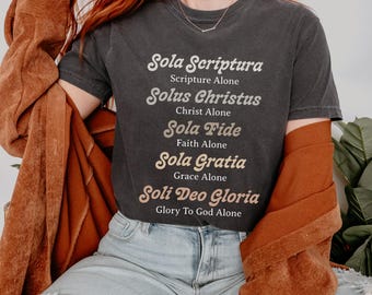 May include: Dark gray t-shirt with the words "Sola Scriptura, Solus Christus, Sola Fide, Sola Gratia, Soli Deo Gloria" printed in a serif font. The shirt is paired with light blue jeans and a brown jacket.