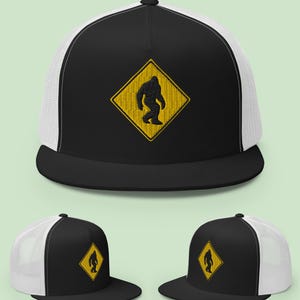Puede incluir: Gorra de camionero negra y blanca con un parche de diamante amarillo con una silueta negra de Bigfoot. La gorra tiene una visera negra y una parte trasera de malla blanca. El texto "Fresh Brim Shack" y "A HAT SHOP" están impresos.