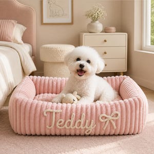 May include: A rectangular, light pink pet bed with a soft, ribbed corduroy texture. The front features the name Teddy embroidered in a cursive script, accompanied by a decorative bow applique. A small, cream-colored plush toy rests inside.