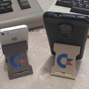 May include: Two cell phones are propped up in a stand with a blue and red logo. The stand is white and the phones are black and silver.