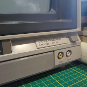 May include: A vintage video monitor with a gray and black exterior. The monitor has a label that reads "VIDEO MONITOR MODEL 1702". The monitor has a power button and two input jacks labeled "VIDEO" and "AUDIO".