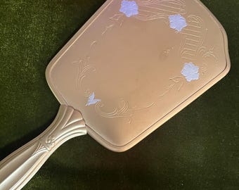 May include: A vintage hand mirror with a light gold finish. The mirror's surface is adorned with delicate floral and scrollwork designs in a soft blue hue. The handle features a ribbed design.