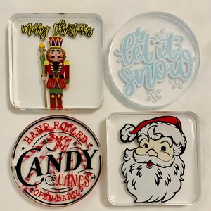 May include: Four clear resin coasters with Christmas-themed designs. One square coaster features a nutcracker soldier with "Merry Christmas" text. Another has "Let it snow" text. A round coaster says "Hand Rolled Candy Canes". A square coaster shows Santa.