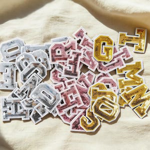 Könnte beinhalten: Eine Sammlung von Buchstabenaufnähern mit Pailletten in Silber, Rosa und Gold. Die Aufnäher sind in verschiedenen Schriftarten und Größen auf einem beigen Stoff verstreut. Die Buchstaben sind weiß umrandet und haben ein glänzendes, strukturiertes Aussehen.