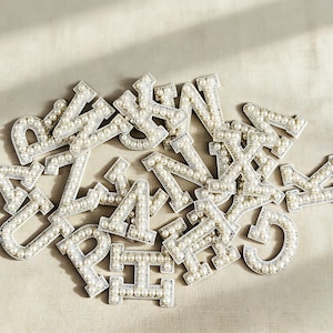 Può includere: Una collezione di toppe a forma di lettera argentate, ciascuna impreziosita da piccole perle bianche rotonde. Le lettere sono sparse su una superficie neutra, creando un elemento decorativo e personalizzato. Ideale per il fai-da-te o per aggiungere stile all'abbigliamento.