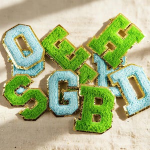 May include: A collection of embroidered fabric letters in various colors. The letters are in shades of light blue and green, with a gold trim. The letters are arranged on a neutral-colored fabric background.