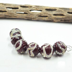 May include: A strand of decorative beads with a deep red base color and swirling white and tan patterns. The beads are round with a ribbed texture. A piece of weathered wood is in the background.