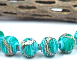 May include: A strand of six round glass beads featuring a vibrant turquoise and white swirled pattern with metallic gold-toned textured accents. The beads are strung on a thin wire, separated by small translucent pink spacer beads at intervals.