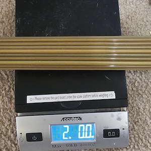 Puede incluir: Una báscula digital que muestra una lectura de 2,00 libras. La báscula es plateada con una base negra y una pantalla digital azul. Un paquete de 12 flechas marrones y tostadas se encuentra en la plataforma de la báscula.