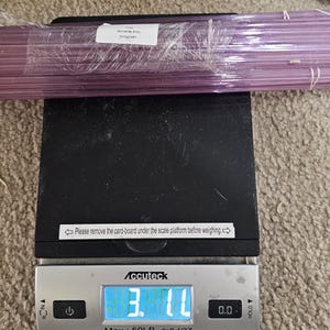 Puede incluir: Una báscula digital con un paquete de alambre morado en la parte superior. La báscula marca 3,10 lb. El paquete está etiquetado con "Wire Gauge 24" y "Made in China".