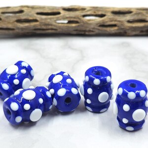 May include: Six cobalt blue glass beads with white polka dots. The beads are cylindrical with a hole running through the center. The beads are scattered on a white surface.