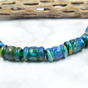May include: A strand of colorful glass beads in shades of blue, green, and yellow. The beads have a swirled pattern and are strung together. A piece of dried wood is in the background.
