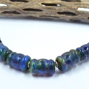 May include: A strand of cylindrical glass beads featuring a glossy finish with swirling patterns of deep blue, emerald green, and hints of purple. The beads are arranged in a curved line, showing a smooth, reflective surface and uniform shape.