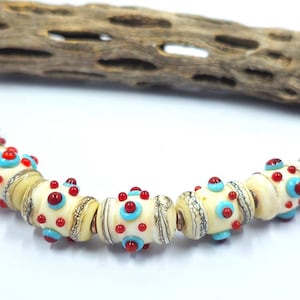May include: A strand of cylindrical glass beads featuring an off-white base color with raised red and light blue dots. Each bead is accented with metallic gold-toned bands and intricate black line detailing, arranged in a curved horizontal line.