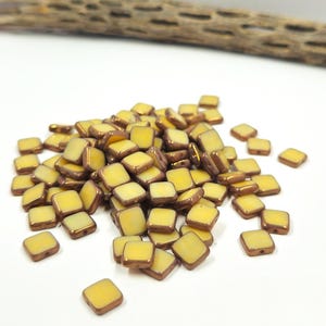 May include: A pile of square beads with a yellow center and a brown border. The beads are scattered on a white surface. The beads are small and appear to be made of glass or a similar material.