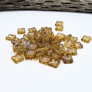 May include: A collection of amber-colored, star-shaped glass beads with a textured, mottled appearance. The beads are scattered on a white surface, with a piece of weathered wood in the background. These beads are ideal for jewelry making and crafting projects.