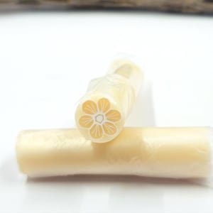May include: Two cylindrical polymer clay canes wrapped in clear plastic film. The end of the upright cane displays a detailed six-petaled flower pattern in light yellow and white, while the horizontal cane is a solid, smooth light yellow.