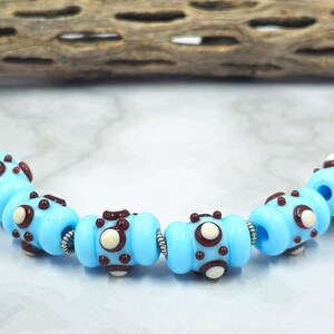 May include: A strand of light blue glass beads with dark brown and white accents. The beads are cylindrical with a series of small, decorative elements. The beads are strung together with silver-colored metal findings.