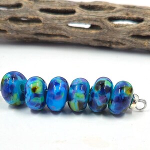 May include: A string of six oval-shaped glass beads in shades of blue, green, and purple. The beads have a marbled appearance and are strung on a silver wire. A piece of driftwood is in the background.