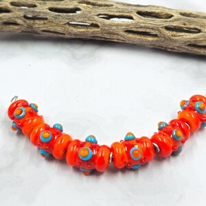 May include: A strand of vibrant orange glass beads with blue and red accents. The beads are round with a slightly flattened shape, strung together with silver-colored wire. The beads are approximately 0.75 inches in diameter, and are arranged in a curved shape.
