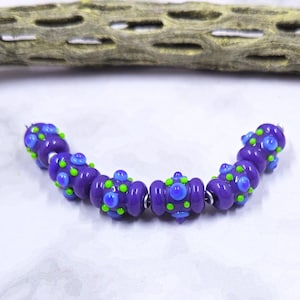 May include: A strand of purple glass beads with blue and green accents. The beads are strung together with silver-colored metal rings. The beads have a rounded shape with raised, colorful details.