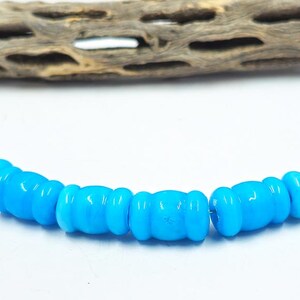 May include: A strand of bright turquoise glass beads. The beads are barrel-shaped with a rounded design. A piece of weathered wood is in the background.