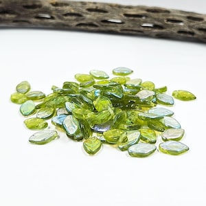 May include: A pile of green leaf-shaped glass beads with iridescent edges. The beads are a mix of light and dark green, with a glossy finish. A piece of brown wood is in the background.