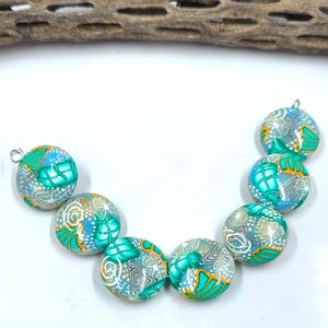 Könnte beinhalten: Eine Sammlung runder, handgefertigter Perlen mit aufwendigen Mustern. Die Perlen zeigen eine Mischung aus türkis, blau, weiß und orangefarbenen Designs. Jede Perle hat eine silberfarbene Schlaufe zum Auffädeln. Die Perlen sind bogenförmig angeordnet.