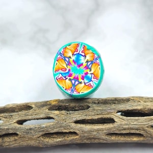 Puede incluir: Una cuenta redonda de arcilla polimérica colorida con un diseño de caleidoscopio. La cuenta tiene un borde turquesa y un patrón central de naranja, azul, rosa y amarillo. La cuenta está sobre un trozo de madera envejecida.