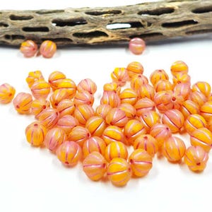 May include: A collection of orange and pink striped glass beads. The beads have a round shape with a ribbed design. The background is white with a piece of wood.